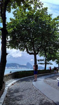 a woman walking down a sidewalk next to trees and the ocean with mountains in the background