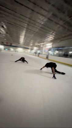 two people are skating on the ice in an indoor rink, one is bending over