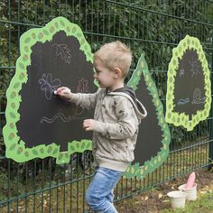 a young boy writing on a chalkboard in front of a fence with other children's drawings