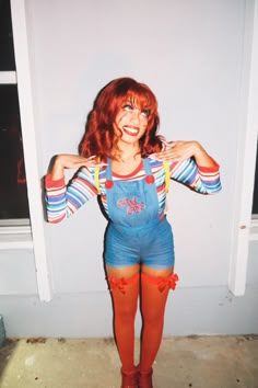 a woman with red hair and blue overalls posing in front of a white wall