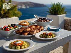 an outdoor table with plates and bowls of food on it, overlooking the water's edge