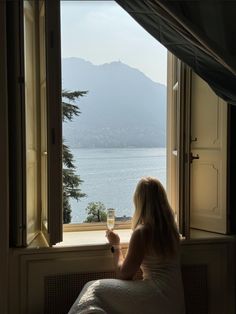 a woman sitting on a window sill looking out at the water and mountains in the distance