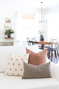 three pillows on a white couch in front of a dining room table with chairs and a potted plant