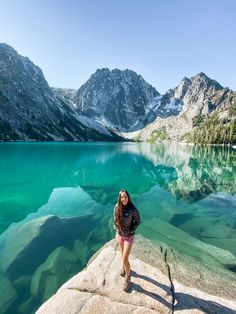 a woman standing on top of a rock next to a lake with mountains in the background