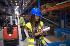 a woman wearing a hard hat and holding a piece of paper while standing in a warehouse