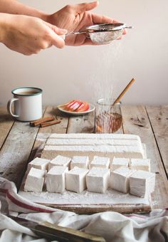 someone sprinkling sugar onto marshmallows on a wooden table with other ingredients