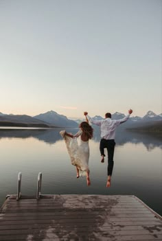 a man and woman jumping into the air from a dock over water with mountains in the background