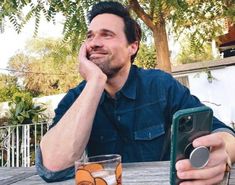 a man sitting at an outdoor table with a cell phone in his hand and two glasses on the table