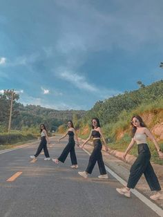 four women in black and white outfits walking across the middle of an empty road with trees on both sides