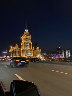 cars driving down the road in front of a large building at night with lights on