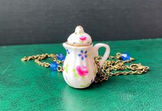 a small tea pot with a chain around it sitting on a green counter top next to a blue and white flowered necklace