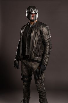 a man dressed in black leather with a helmet and goggles, standing against a gray background