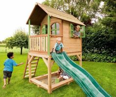 two children playing in a wooden play house with a slide and green grass field behind them