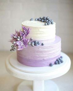 a three layer cake with purple frosting and flowers on the top, sitting on a white pedestal