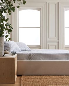 a bed sitting in front of two windows next to a wooden table with a potted plant on it