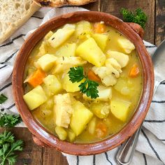 a bowl filled with chicken and potatoes next to bread on a wooden table, surrounded by fresh parsley