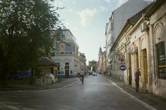 two people walking down the street in front of buildings and trees on either side of the road