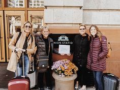 three women standing next to each other with luggage and flowers in front of a building