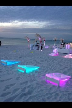 an outdoor event set up on the beach at night with colorful lights and chairs in the sand