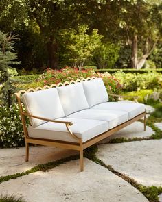 a white couch sitting on top of a stone walkway next to flowers and trees in the background