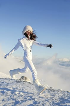 a woman in white snowsuit and hat on top of a snowy hill with her arms outstretched