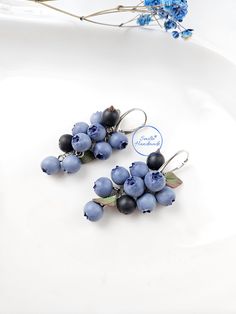 two pairs of earrings with blue berries on them sitting on a white plate next to flowers