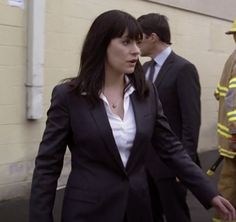 a woman in a suit and tie walking down the street with fire fighters behind her