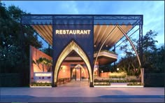 the entrance to a restaurant at dusk with trees and bushes in the foreground, lit up by lights