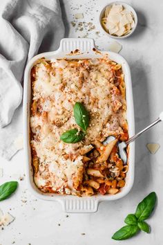 a white casserole dish filled with pasta and sauce, garnished with fresh basil