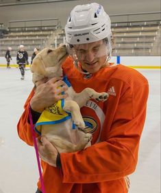 a man in an orange jersey holding a dog on a hockey rink with other players