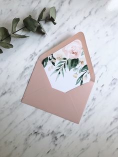 an envelope with flowers on it sitting next to a rose and eucalyptus leaf arrangement in front of a marble background