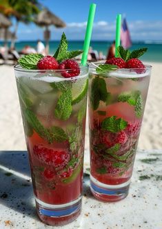 two glasses filled with raspberries and minty drinks on a table at the beach