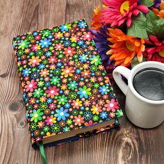 a colorful flowered notebook next to a cup of coffee on a wooden table with flowers