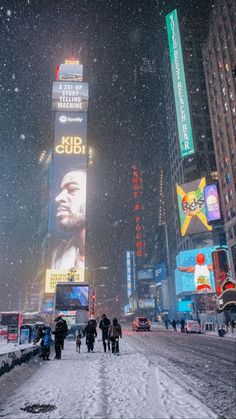 people walking in the snow on a city street with billboards lit up at night