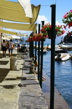 people are walking on the sidewalk next to water with boats and flowers in pots hanging above them