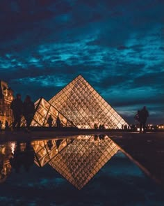 people are standing in front of the glass pyramid at night, with lights reflecting on the water