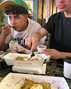 two young men sitting at a table eating food from paper plates and cups with spoons