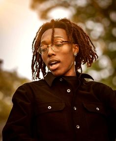 a woman with glasses and dreadlocks standing in front of trees wearing a black shirt