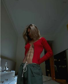 a woman with no shirt standing on a bed wearing green pants and a red sweater