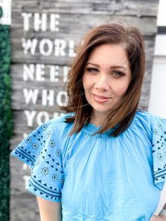 Brunette woman standing in front of a wooden palette sign that reads "The world needs who you were made to be." She is wearing a bright blue top with embroidery on the bell cap sleeves, a mock. neck with mini ruffles and mini pleats. Her hair is windblown. Sleeve Embroidery, White Belt, Cute Sandals