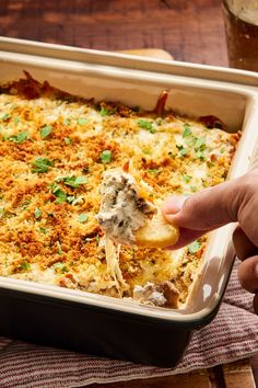 a person dipping some food into a casserole dish with meat and cheese on top