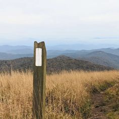 there is a wooden sign in the middle of a grassy field with mountains in the background