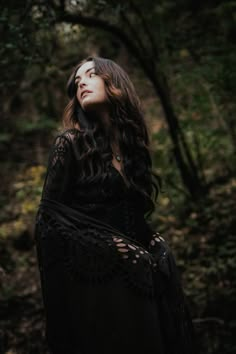 a woman with long hair standing in the woods wearing a black dress and shawl