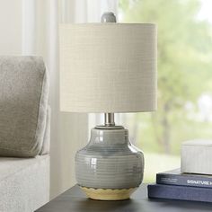 a table with a lamp on it next to a book and a couch in front of a window