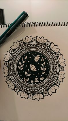 a black and white drawing on top of a notebook next to a pen, with a flower design in the middle