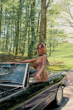 a woman standing on the hood of a black car in front of trees and grass