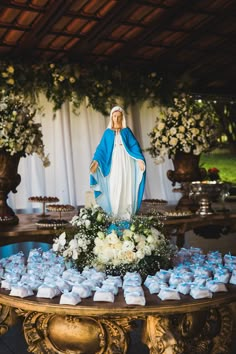 a statue of the virgin mary surrounded by blue and white flowers on top of a table