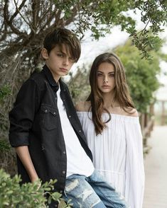 two young people standing next to each other near some bushes and trees, one is wearing a white shirt