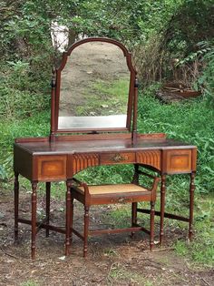 an antique dressing table with mirror and stool in the middle of some grass, next to trees