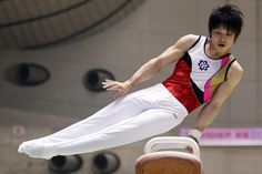 a man is doing tricks on the balance beam in an indoor gymnastics arena while wearing white pants and a red tank top